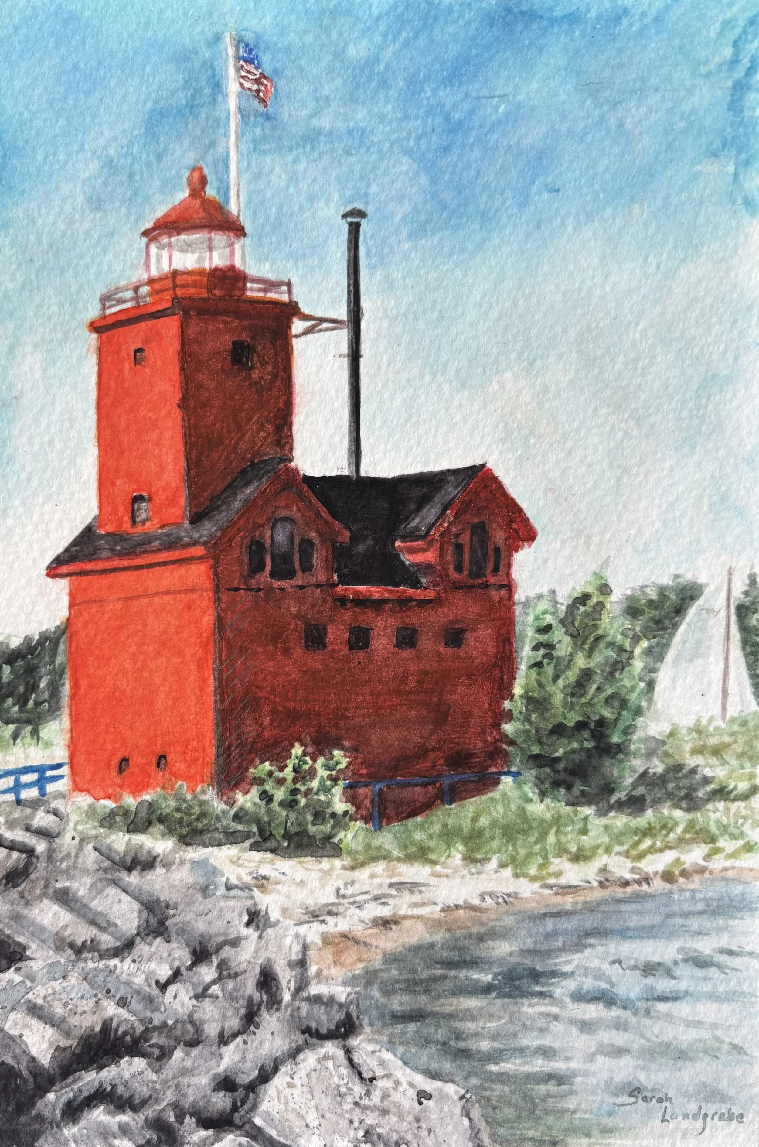 Watercolor painting of a red lighthouse next to a rocky shoreline and water with green trees in the background under a blue sky.