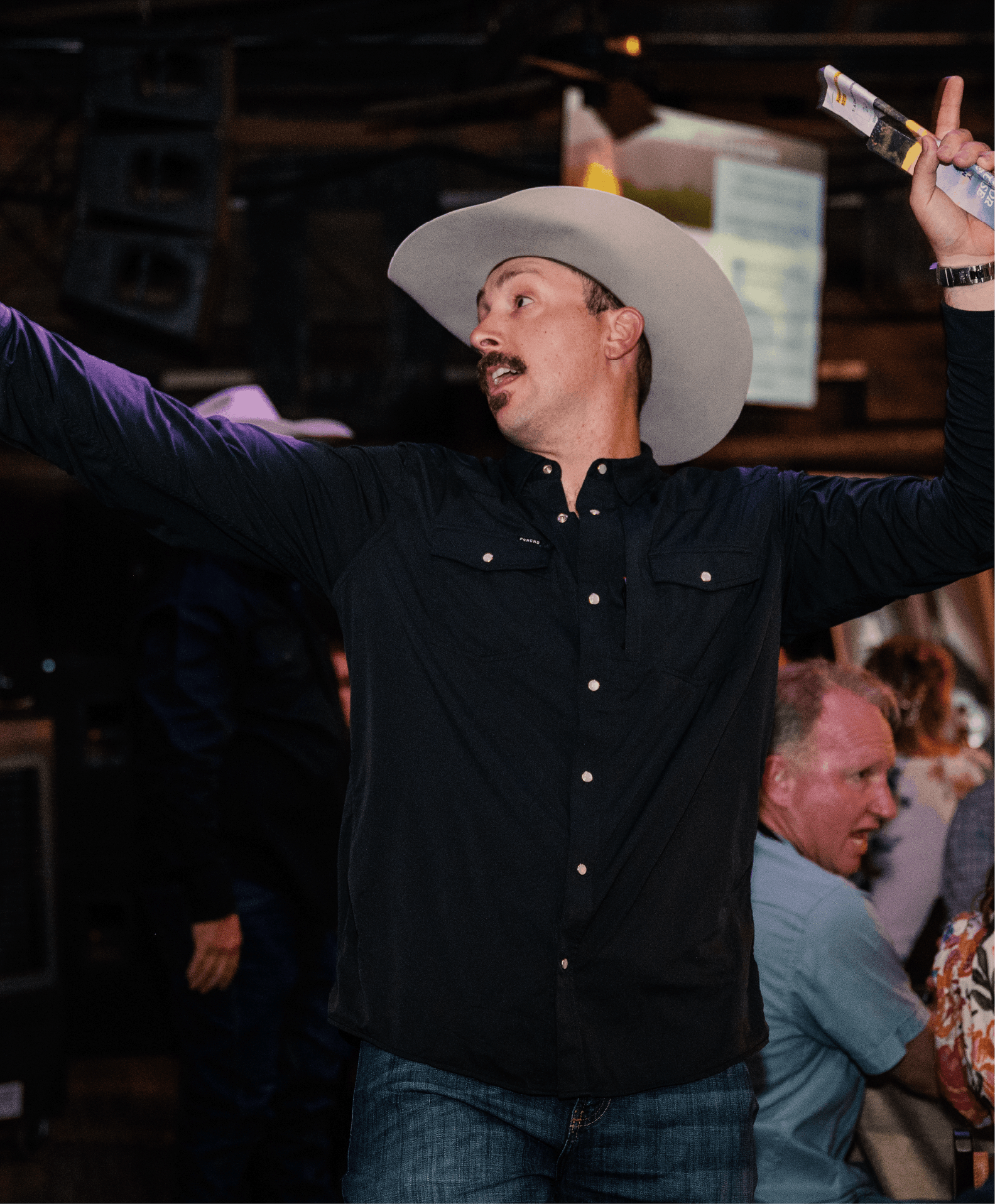 A man wearing a large cowboy hat and black shirt appears to be singing or speaking passionately, with his arms raised, at a social event or party.