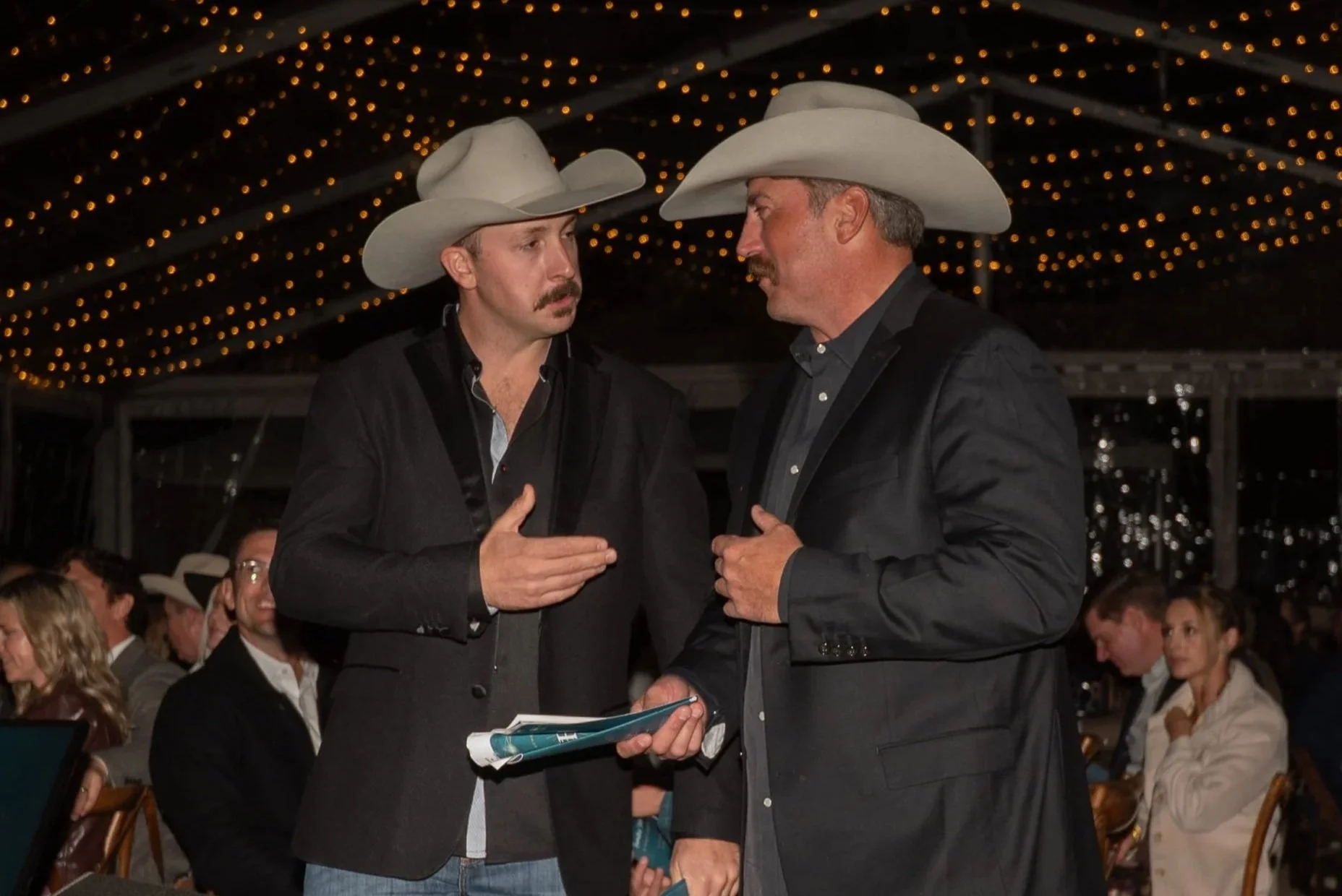 Two men wearing cowboy hats and dark jackets are engaged in a conversation at an event with string lights overhead. They are standing close, gesturing with their hands, and appear to be discussing something seriously. Other people are seated in the background.