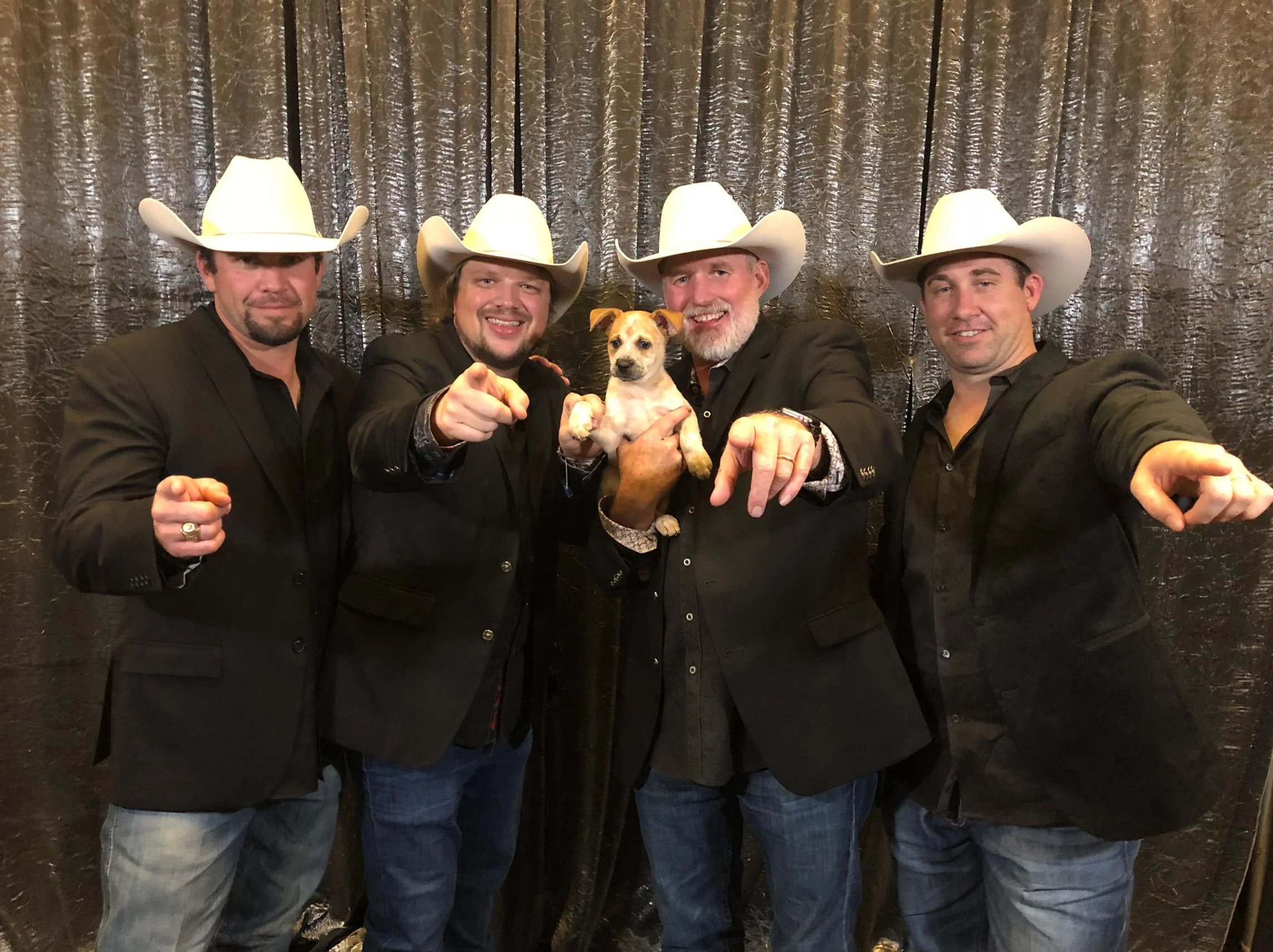 Four men wearing black suits and white cowboy hats, posing with a dog in front of a black, textured backdrop. The men are smiling and pointing at the camera.