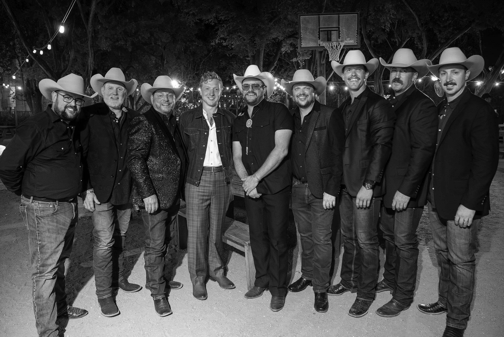 Group of ten men dressed in cowboy hats and semi-formal attire, standing outdoors at night with string lights and a basketball hoop in the background.