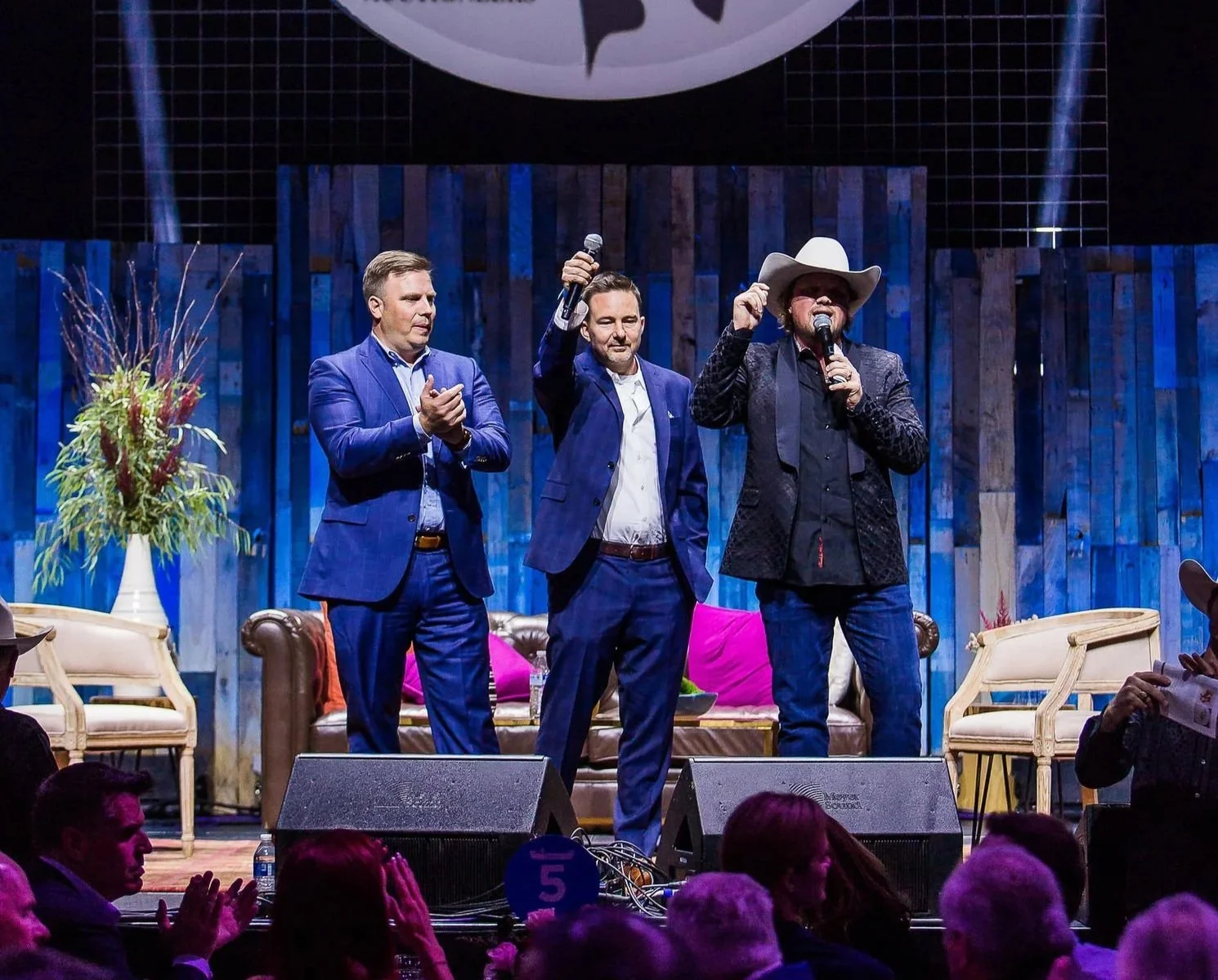 Three men on stage at the Cowboy Auctioneer event, with a large sign displaying 'Cowboy Auctioneer by Heath Hale Auctioneers' and photos of children in the background, a woman in a cowboy hat speaking, and an audience clapping.