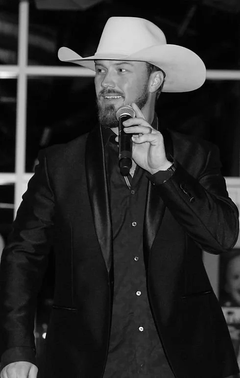 A man wearing a large cowboy hat, black suit, and shirt, holding a microphone and speaking at an event.