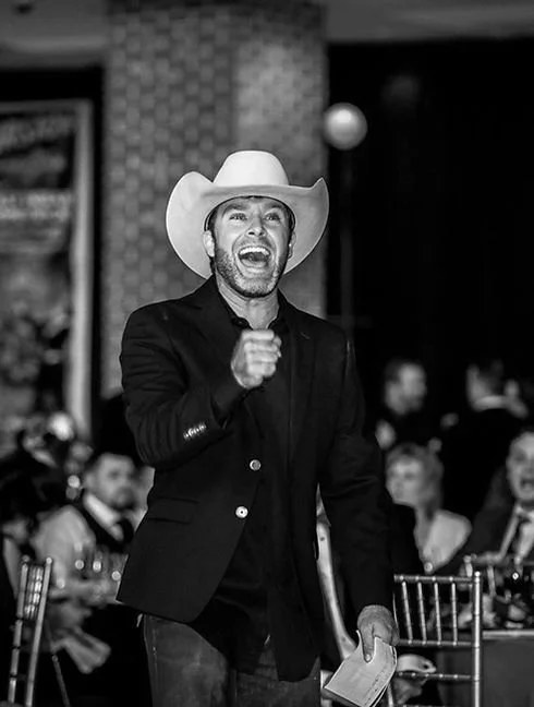 Man wearing a cowboy hat, black blazer, and shirt, smiling and gesturing with one hand at an event with seated guests and a patterned wall in the background.