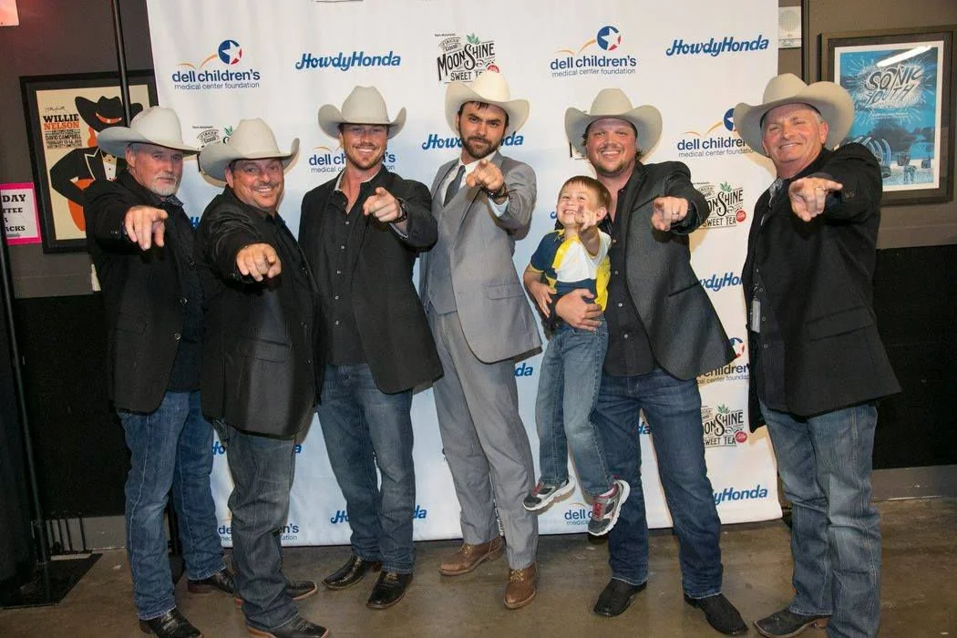 Six men and a child wearing cowboy hats and jackets, pointing towards the camera, standing in front of a patriotic-themed backdrop with logos, including 'Howd Honda,' 'Dell Children's,' and 'Moonshine Sweet Tea'; the man in the middle is dressed in a