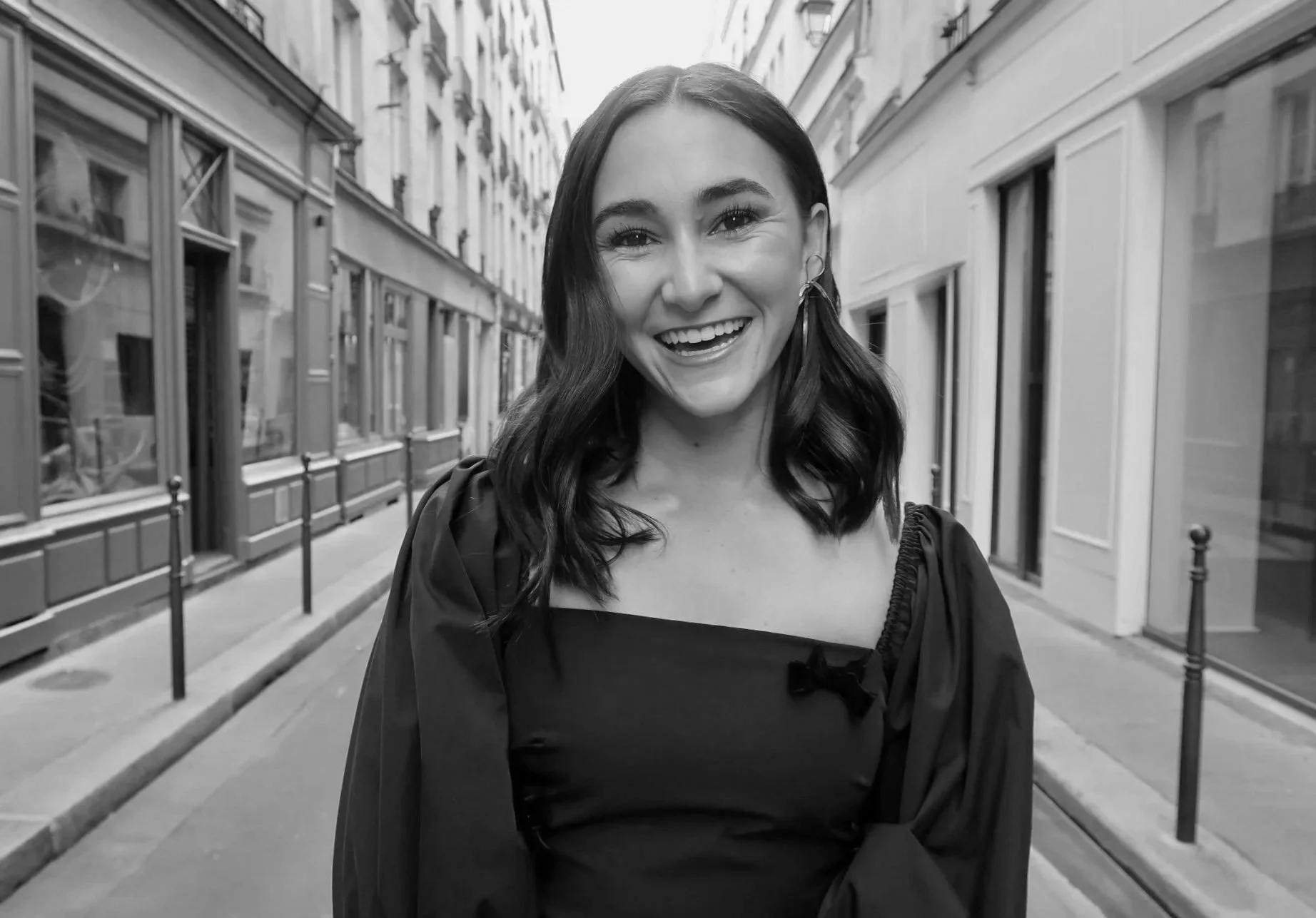 Black and white photo of a young woman smiling on a city street, wearing a dress with puffed sleeves, with buildings lining both sides of the street.