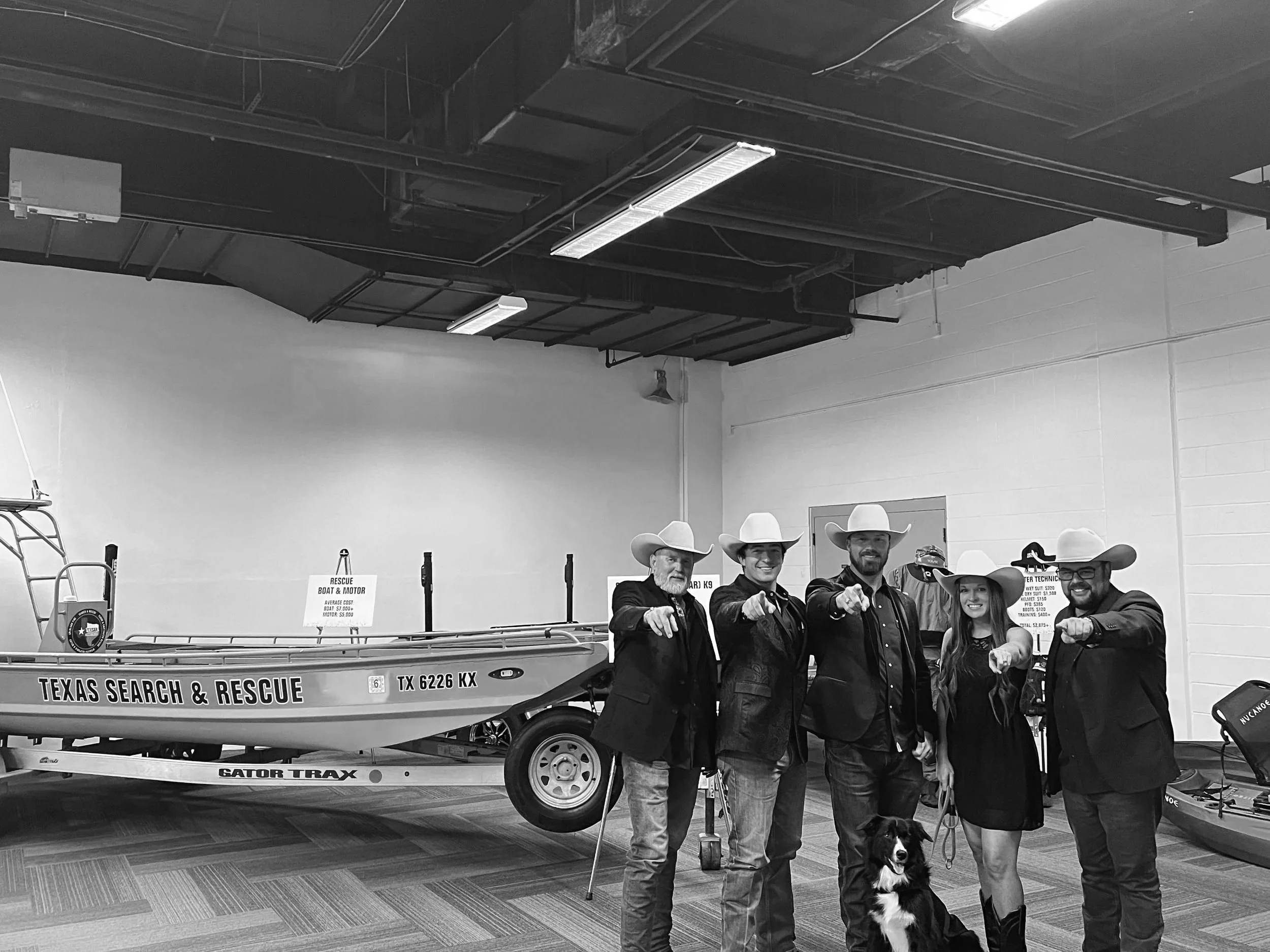 Group of five people wearing cowboy hats, standing inside a building with rescue boats and equipment, pointing at the camera.