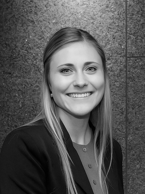 A smiling young woman with long blonde hair, dressed in professional attire, standing in front of a textured stone wall.