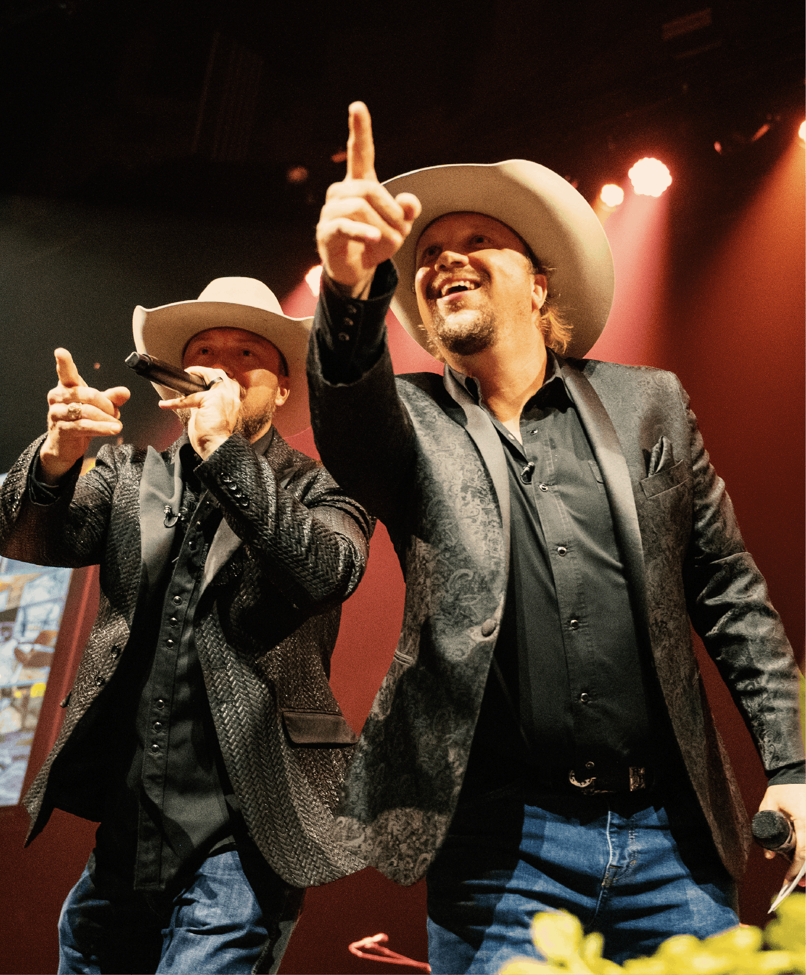 Two men wearing cowboy hats and black suits performing on stage, with one holding a microphone and the other pointing upward, in front of a red background with stage lights.