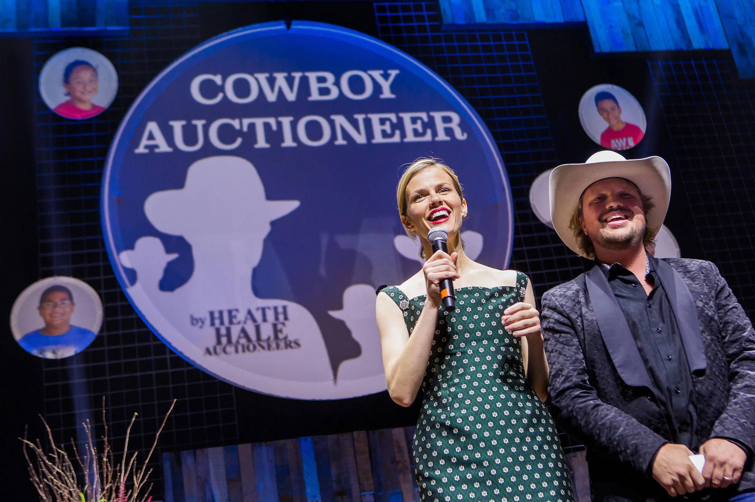 A woman in a green dress holding a microphone and smiling next to a man in cowboy attire with a large hat. Behind them is a sign that reads "Cowboy Auctioneer" with additional text and images of children in circles.