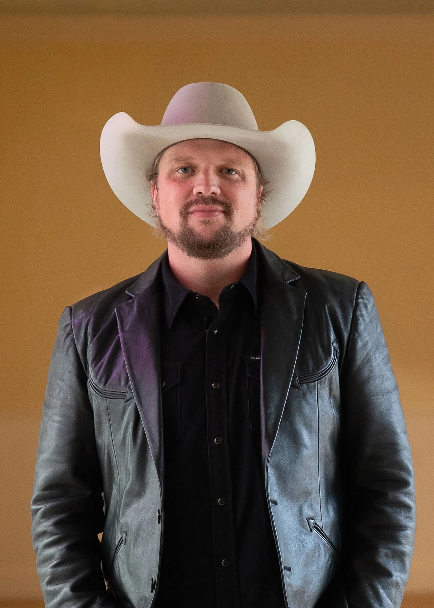 A man wearing a white cowboy hat, a black leather jacket, and a black shirt standing against a tan background.