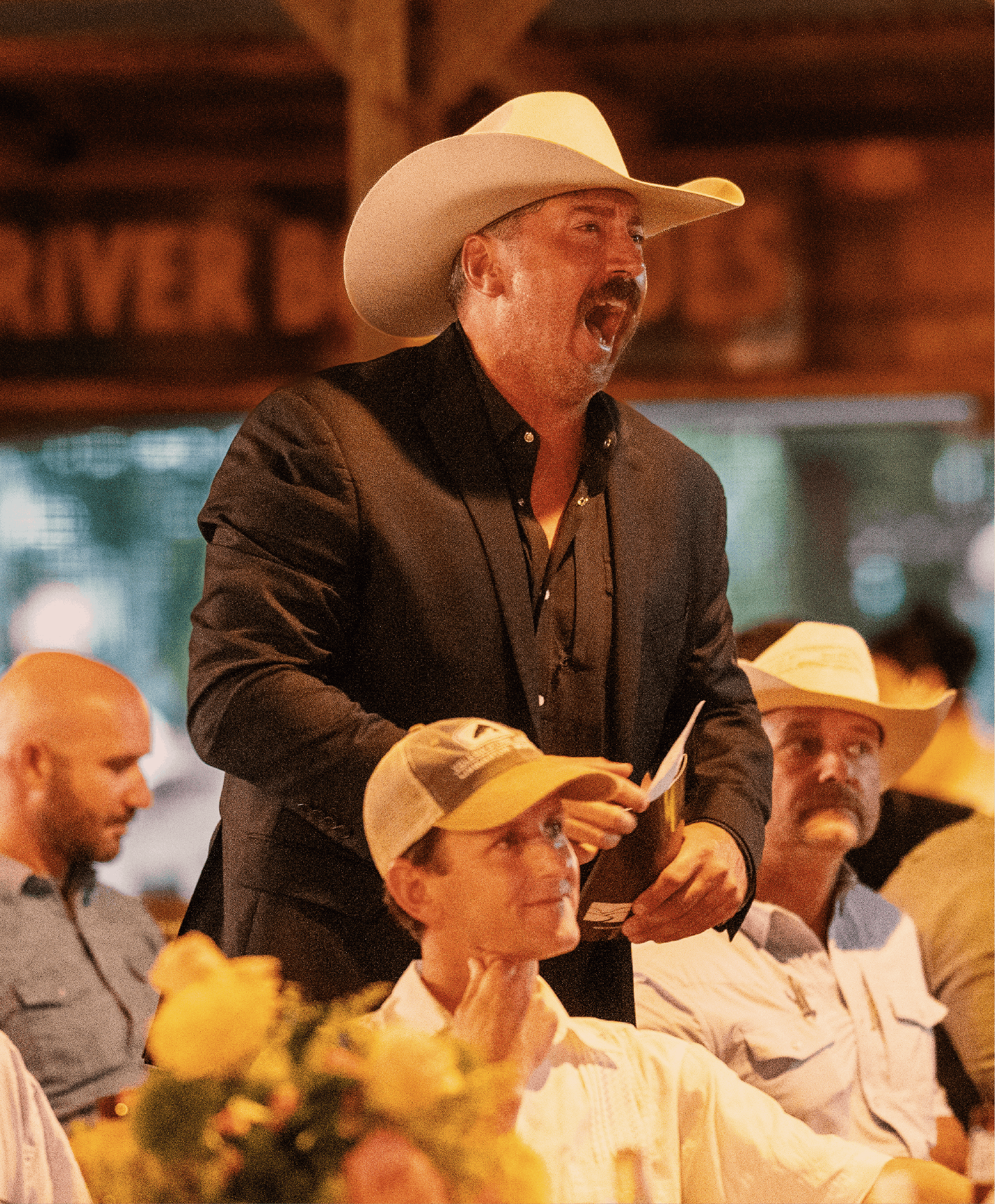 A man wearing a white cowboy hat and black suit passionately speaking or singing at an event, with people in cowboy hats and casual clothing seated around him, indoors with warm lighting.