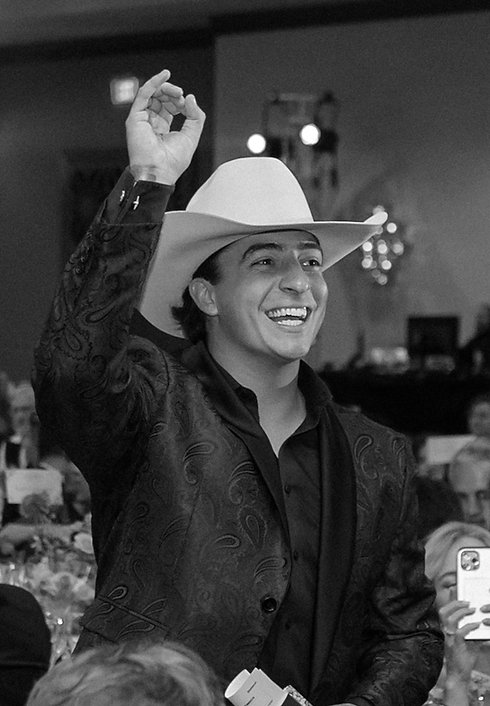 A man wearing a cowboy hat and a dark patterned jacket, smiling and raising his hand at an indoor social event.