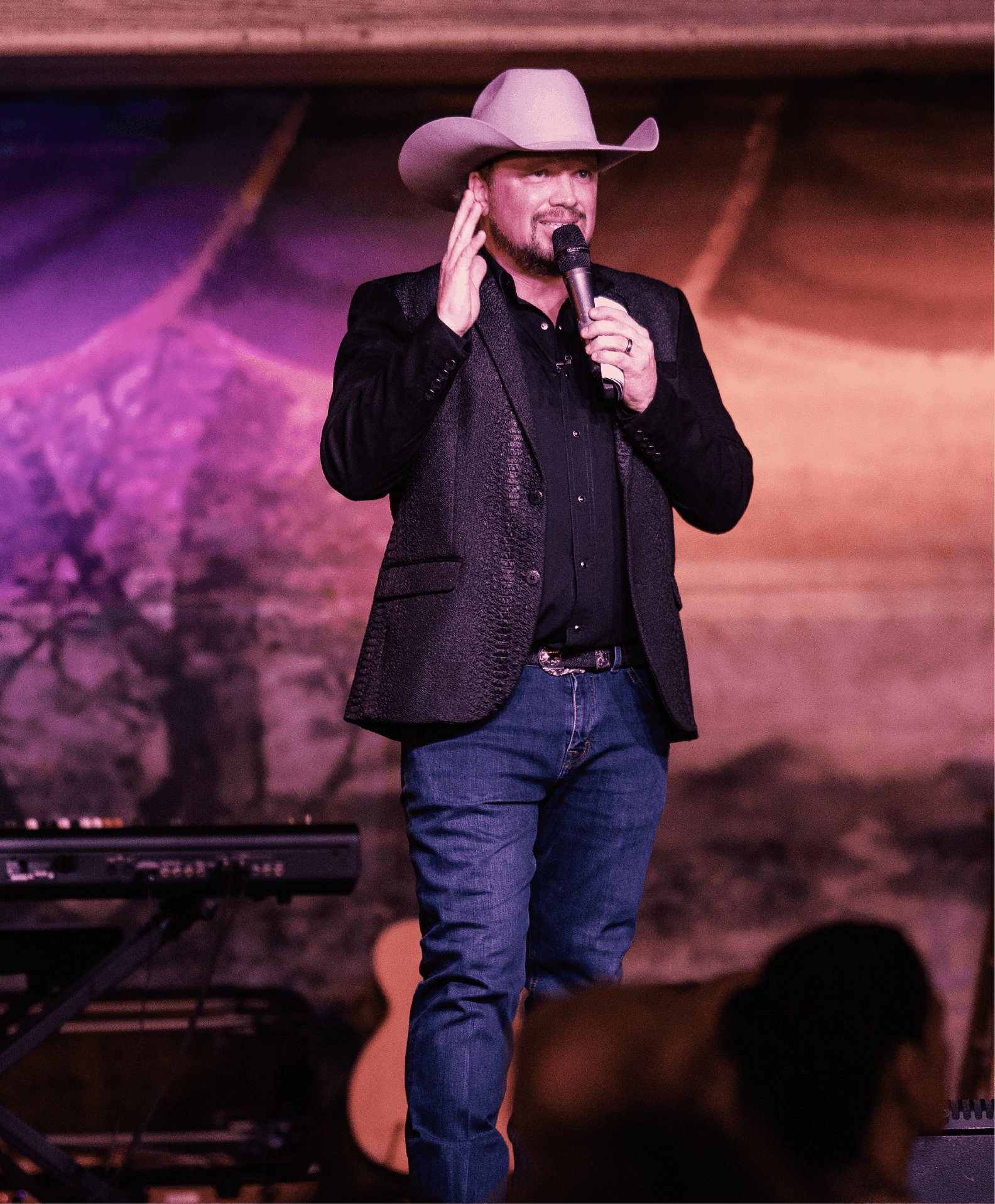 A man wearing a cowboy hat and black blazer, holding a microphone, standing on stage in front of a backdrop with a mountain and trees, with musical instruments nearby.