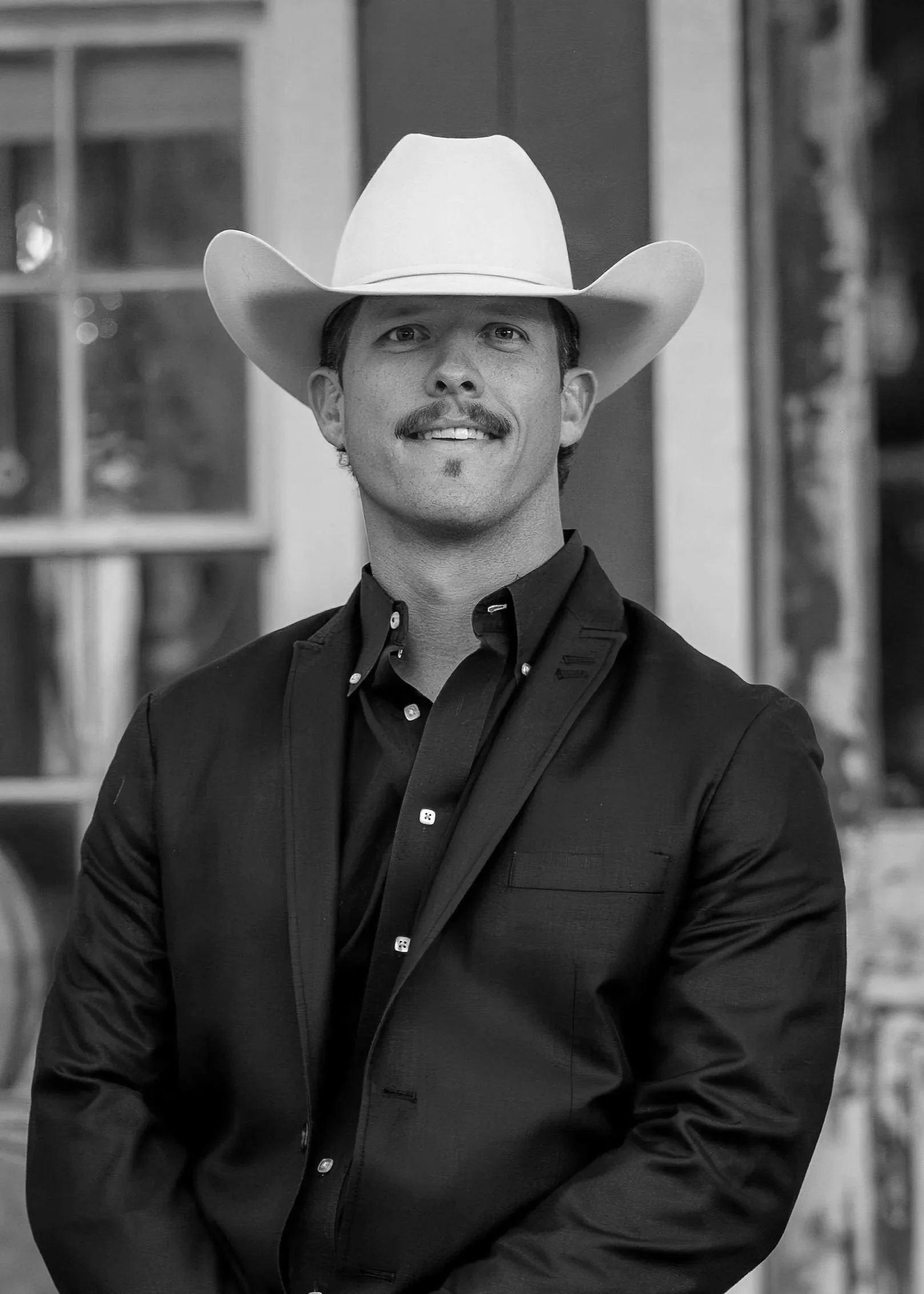 A man wearing a cowboy hat and a dark blazer, smiling at the camera, with a blurred background.