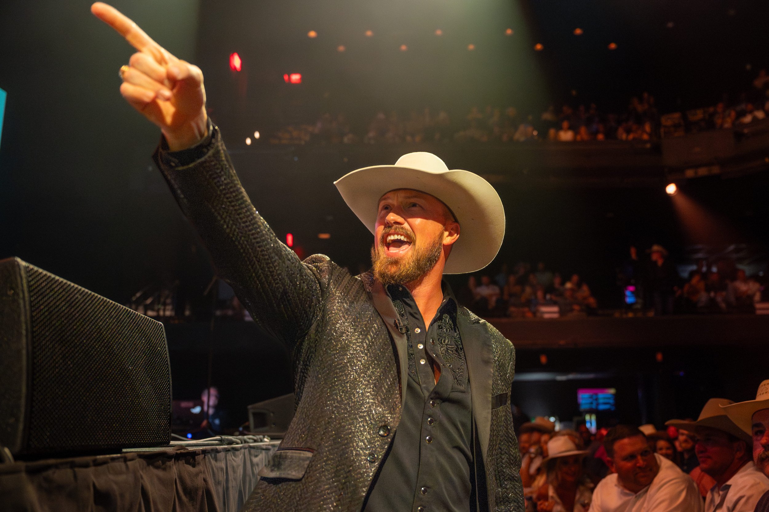 A man wearing a white cowboy hat, black shirt, and shiny black jacket is on stage at an event, smiling and pointing upwards. The background shows an audience seated in a theater.