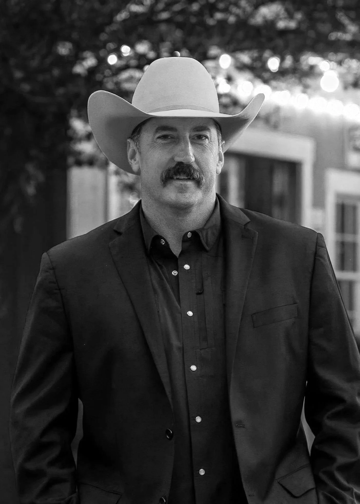 A man with a mustache wearing a cowboy hat and a dark suit jacket, standing outdoors with blurred trees and buildings in the background.