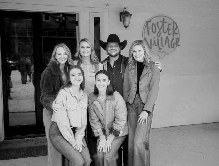 Group of six people, five women and one man wearing a cowboy hat, standing outside a building with a sign that says 'Foster Village'