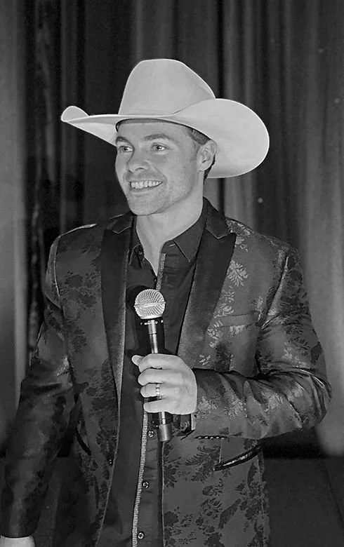 A man wearing a cowboy hat and a patterned suit, smiling, holding a microphone on stage with a curtain background.