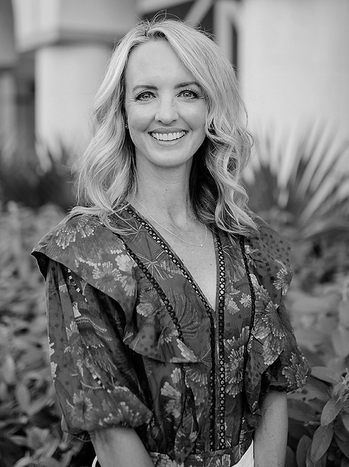 A smiling woman with wavy blonde hair, wearing a patterned dress, standing outdoors with tropical plants and a building in the background.