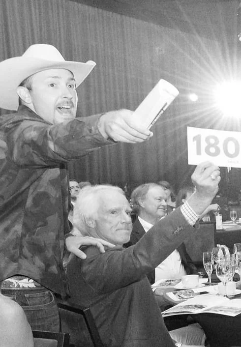 A man wearing a cowboy hat standing and holding a book, while an older man seated next to him holds up a bid number 180 at an auction or event.
