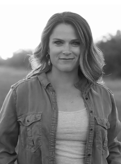 Black and white portrait of a woman with shoulder-length wavy hair, wearing hoop earrings, a casual shirt, and a jacket, standing outdoors with blurred trees behind her.