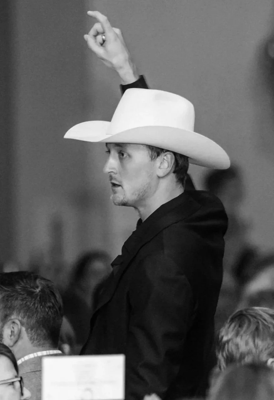A man in profile wearing a cowboy hat and black jacket, raising his right hand while looking at something in front of him.