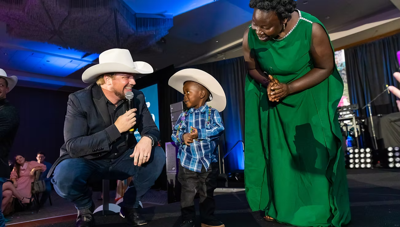 A man wearing a cowboy hat, black jacket, and jeans kneeling and speaking into a microphone, smiling at a young boy in a cowboy hat and plaid shirt, who is standing next to a woman in a green dress on stage at an event.