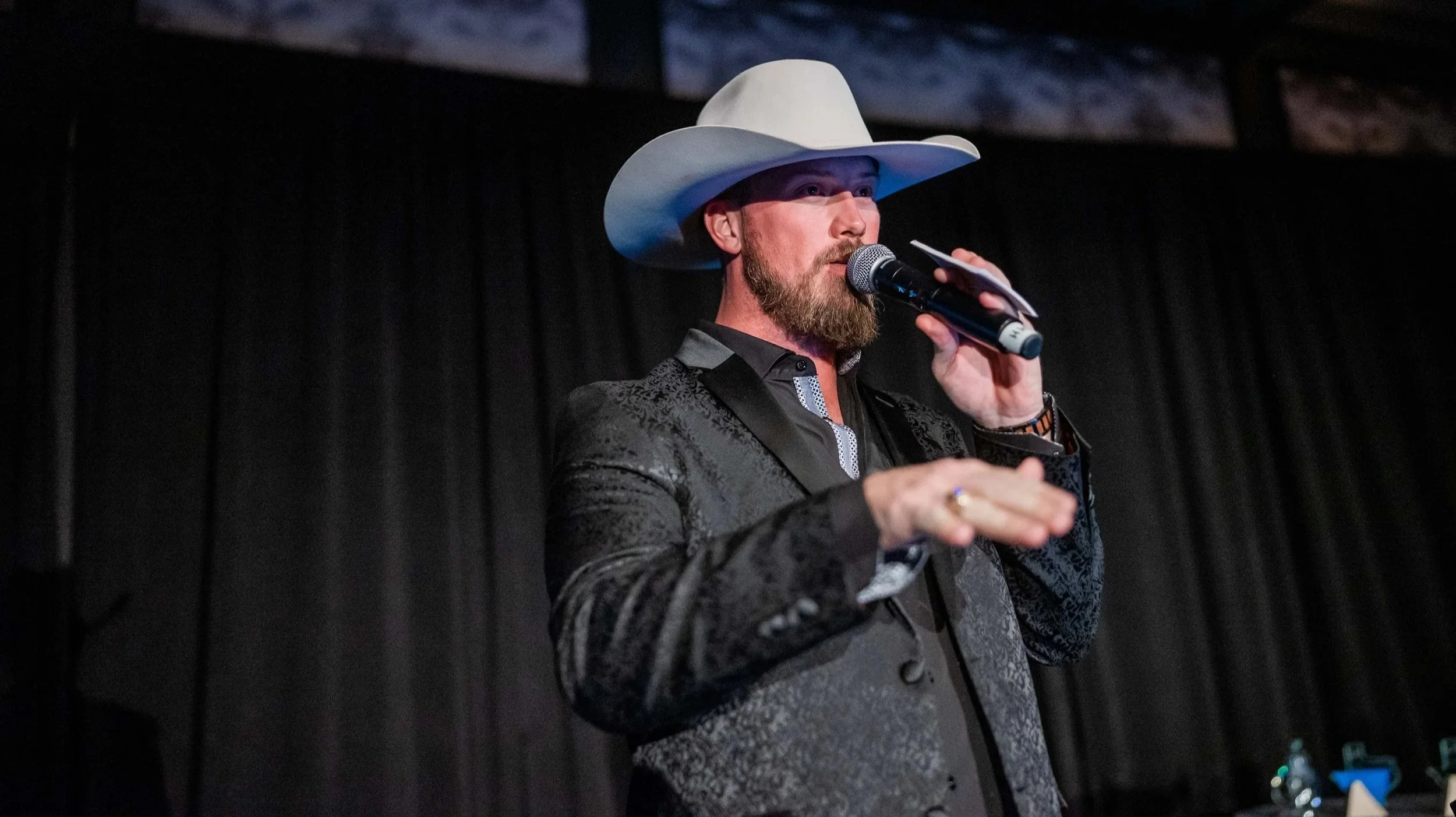 A man with a beard wearing a white cowboy hat, black patterned blazer, and black shirt holding a microphone and speaking on stage against a black curtain backdrop.