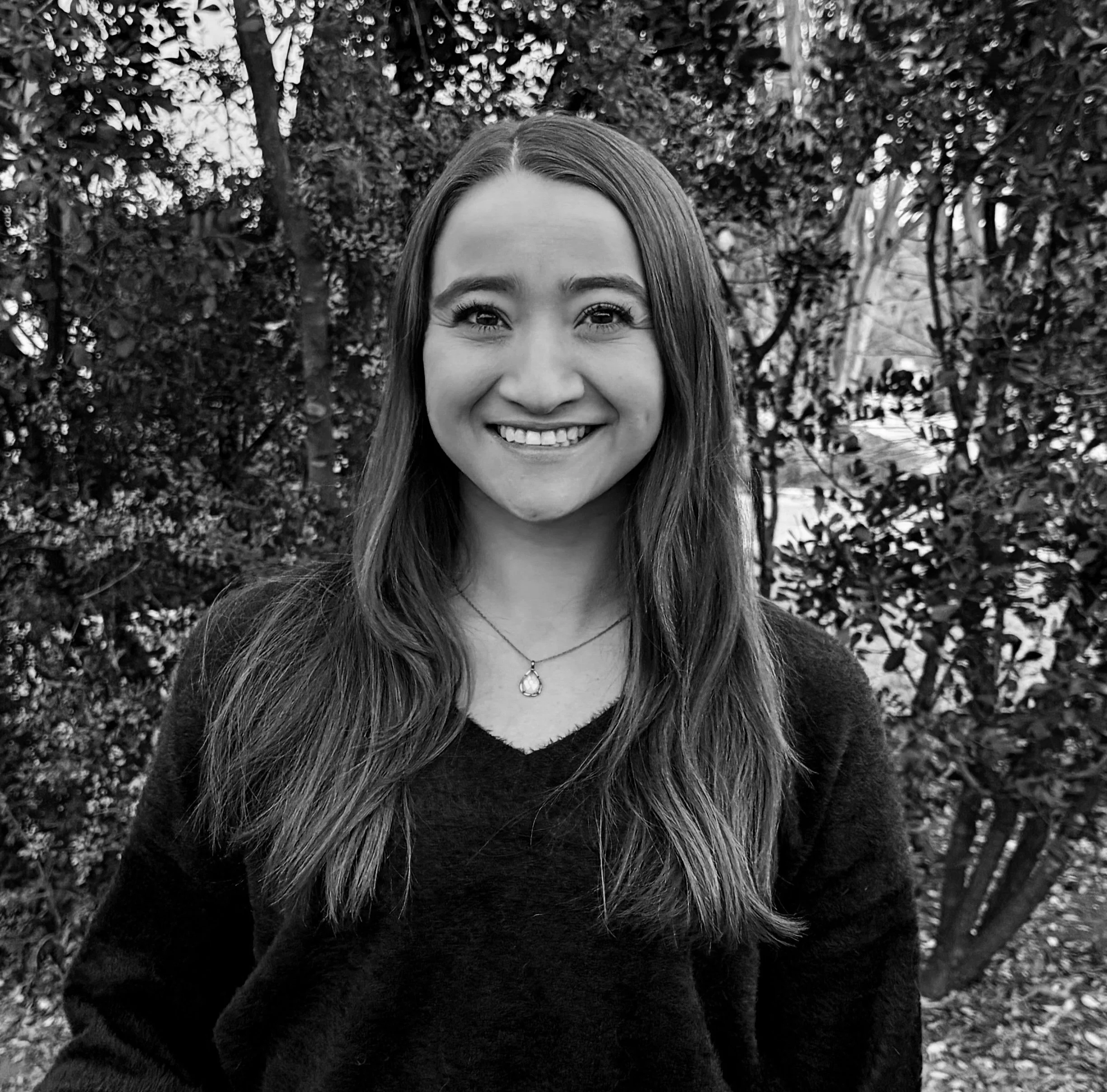 A woman with long hair smiling outdoors, wearing a dark sweater and a necklace, with trees and foliage in the background.