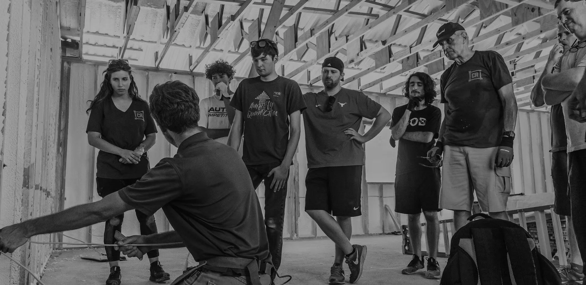 A group of young adults and instructors in a construction or training workshop, observing a person demonstrating something with a tool in a partially built structure.