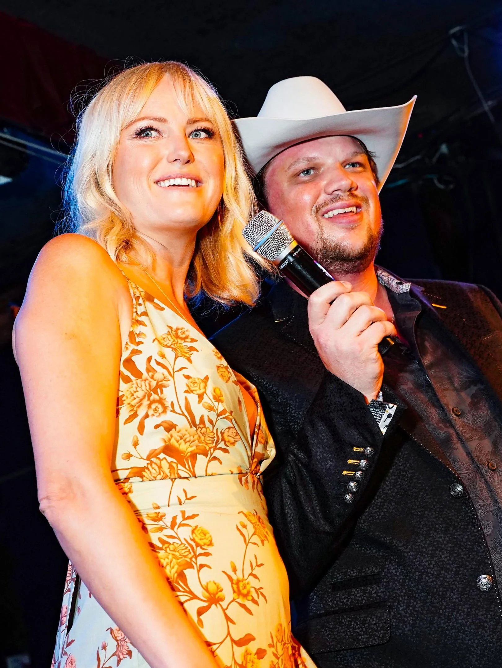A woman with blonde hair wearing a yellow floral dress and a man with a white cowboy hat holding a microphone, both smiling on stage.