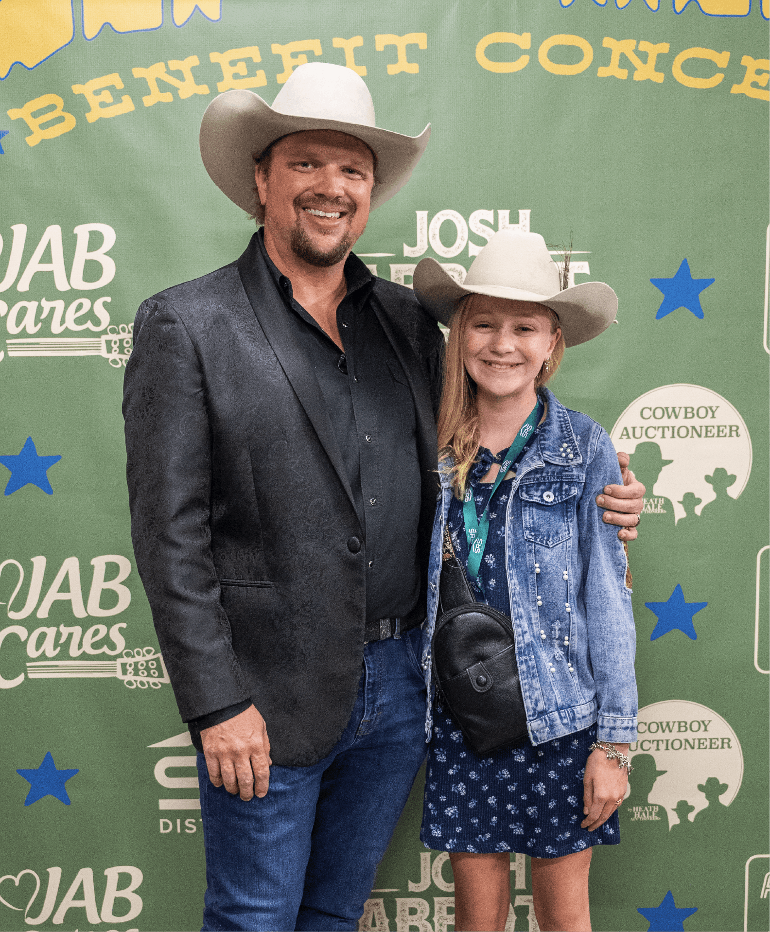 A man and girl dressed in cowboy hats and denim jackets standing together and smiling at the camera at a Western-themed event with a green backdrop featuring country music references and blue star decorations.