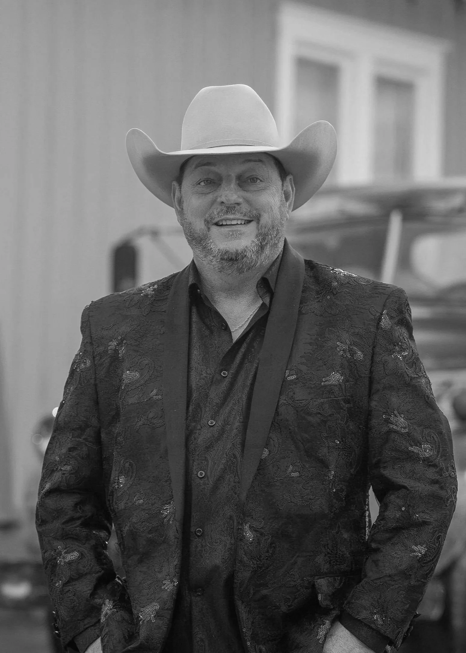 A smiling man wearing a cowboy hat and a dark patterned blazer.