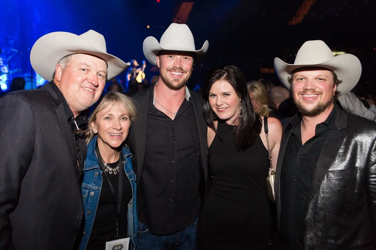 Five people at a concert or event, three men and two women, wearing cowboy hats and dark clothing, smiling for the camera.