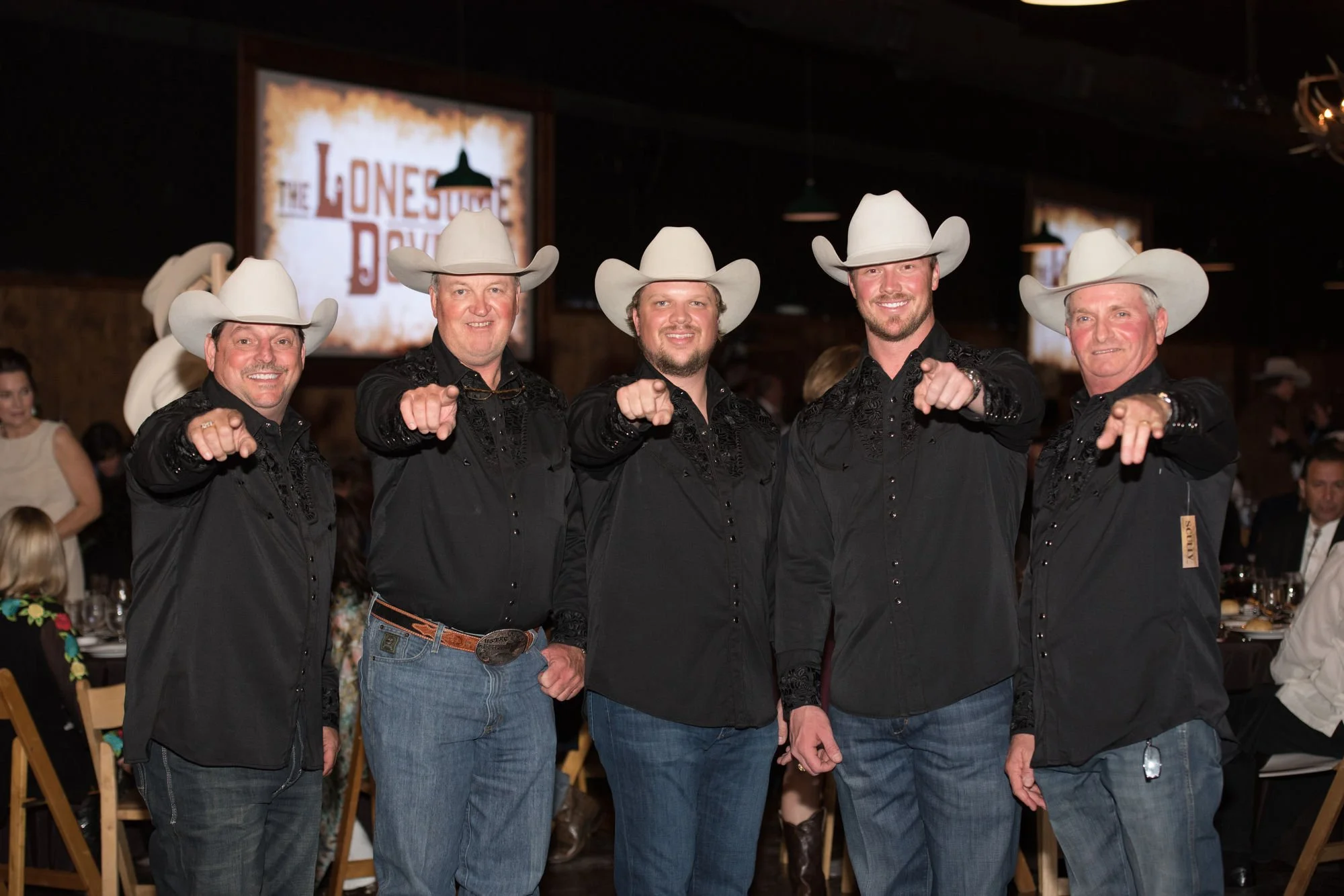 Five men dressed in black shirts and cowboy hats, pointing at the camera at an event, with a sign in the background reading 'The Lonesome Draw.'
