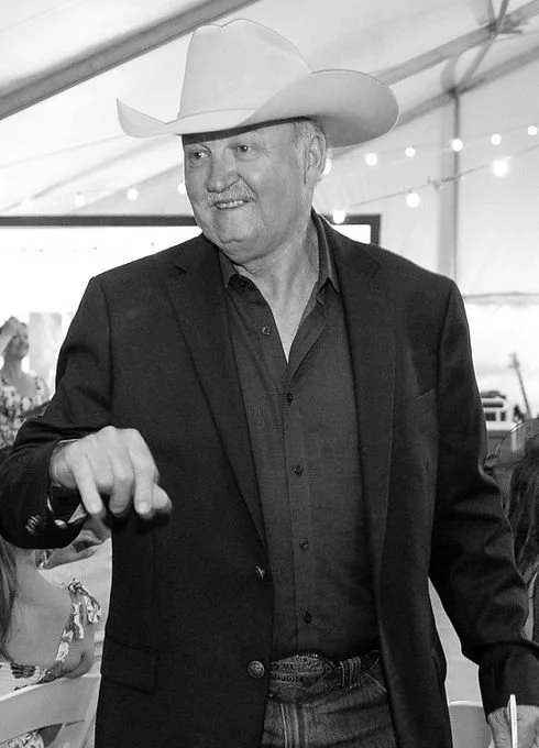 Man in a cowboy hat and dark suit smiling at a social event in a tent.