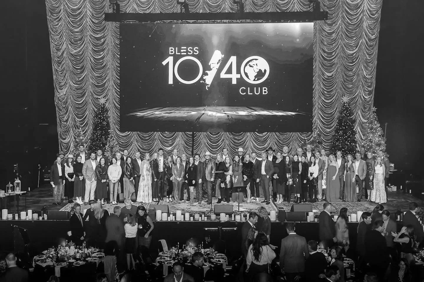 A large group of people on stage during a celebration event with decorated Christmas trees and a big screen displaying 'Bless 10/40 Club' in the background. The setting appears festive and formal.