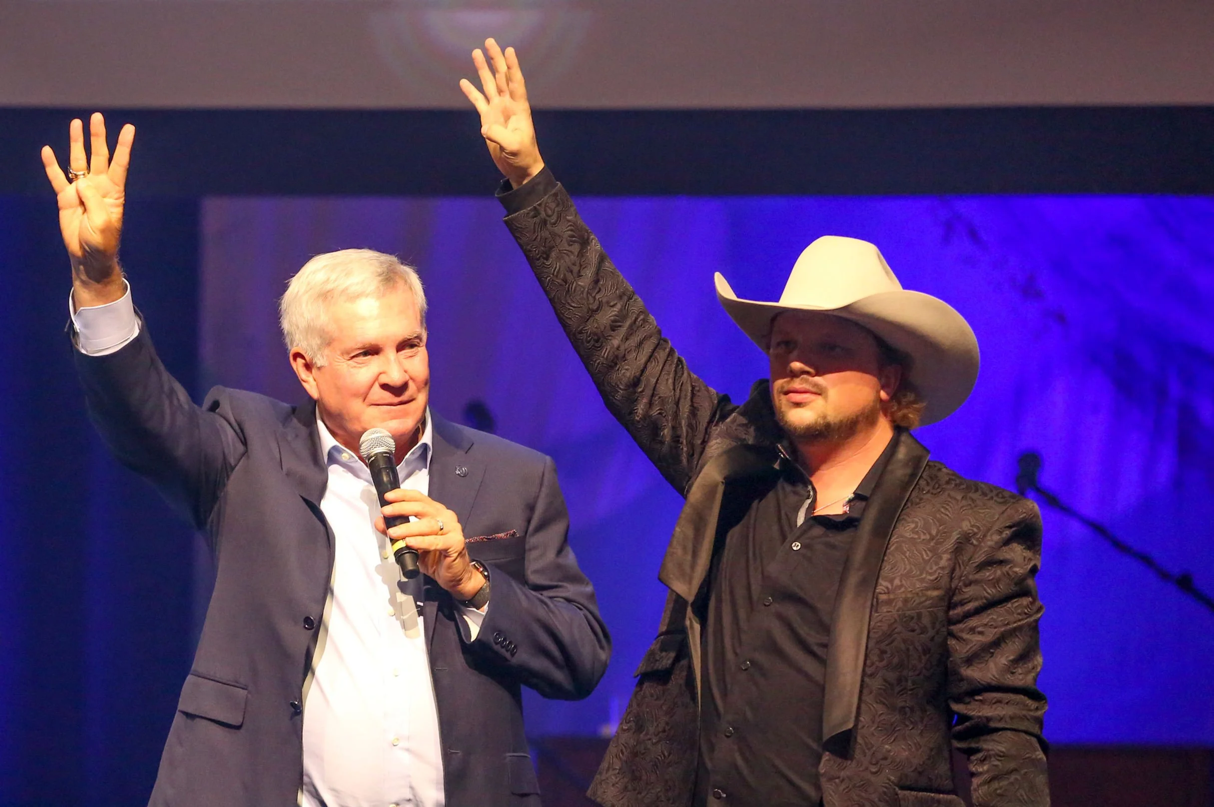 Two men on stage, one in a suit holding a microphone and the other in a cowboy hat and dark patterned jacket, raising their hands in the air.