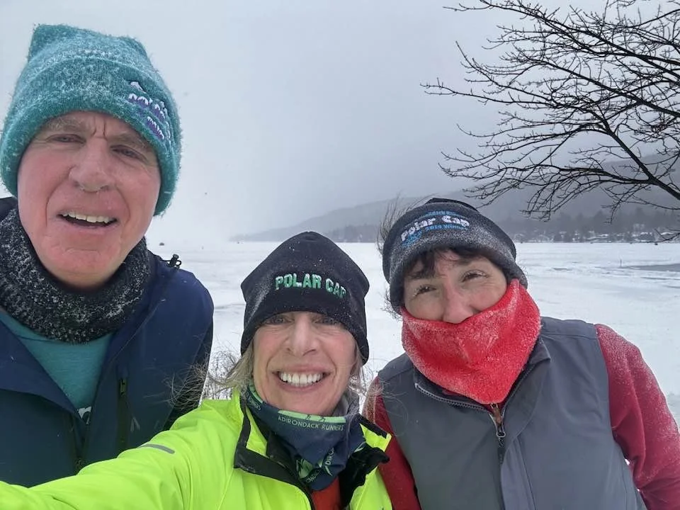 Three people taking a selfie outdoors in winter, dressed warmly with snow and a frozen landscape in the background, and smiling.