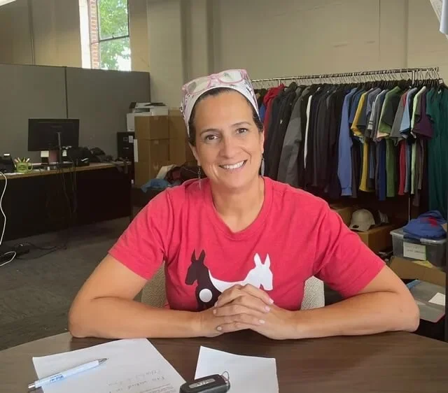 Woman smiling and sitting at a desk with papers, a pen, and a key fob, in a room with a clothing rack and computer equipment.