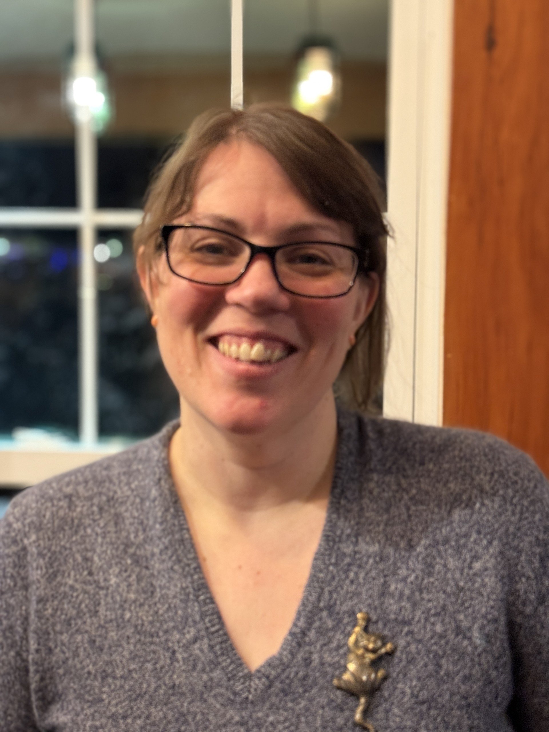 A smiling woman with short brown hair, glasses, and a gray sweater, sitting indoors near a window with a wooden frame. She is wearing a brooch in the shape of a frog.