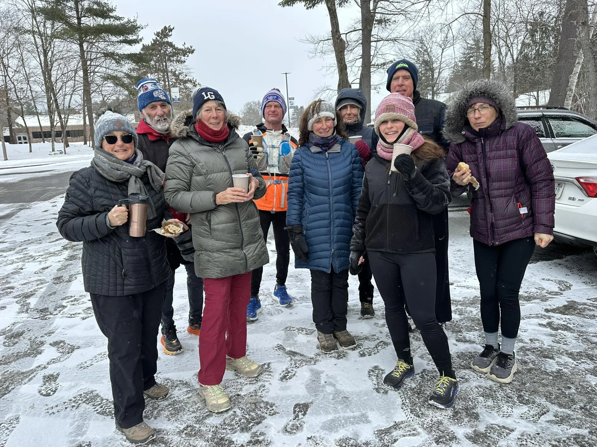 A group of people bundled in winter clothing, standing outdoors on a snowy day, smiling and holding coffee cups and snacks, with cars and trees in the background.