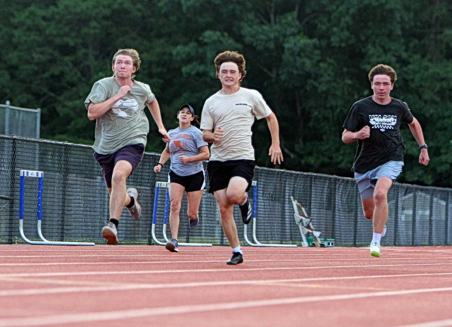 Four young people running on a track during daytime with a green wooded background.