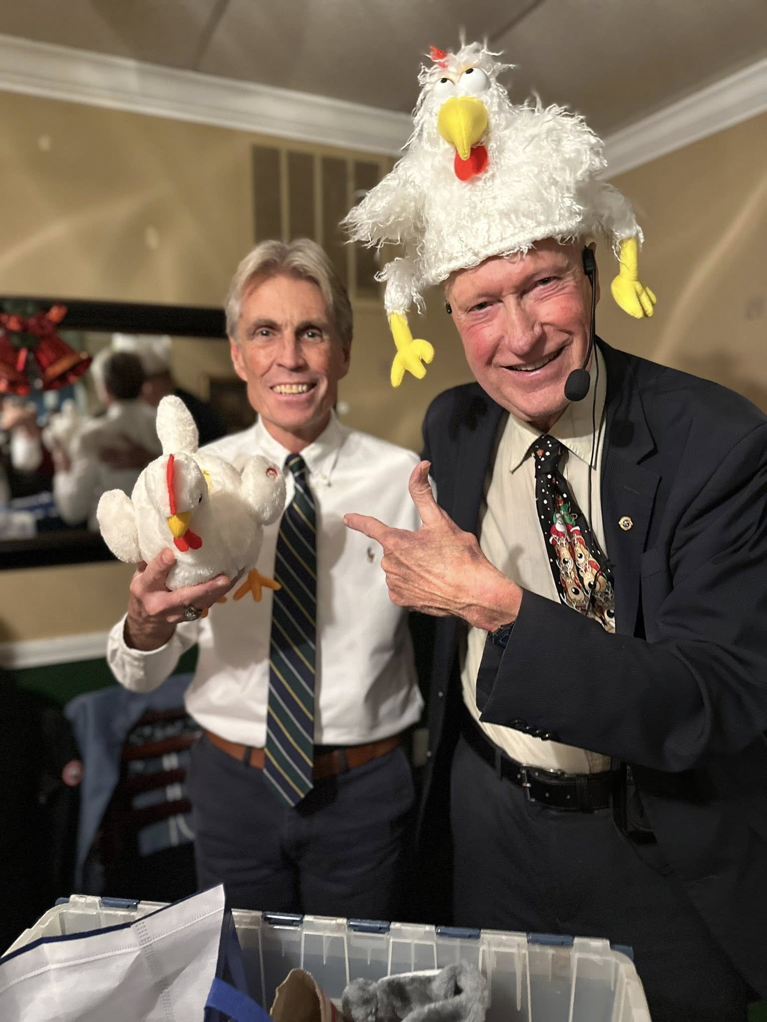 Two men in formal attire posing with playful chicken-themed puppets, one wearing a chicken hat, in an indoor setting.