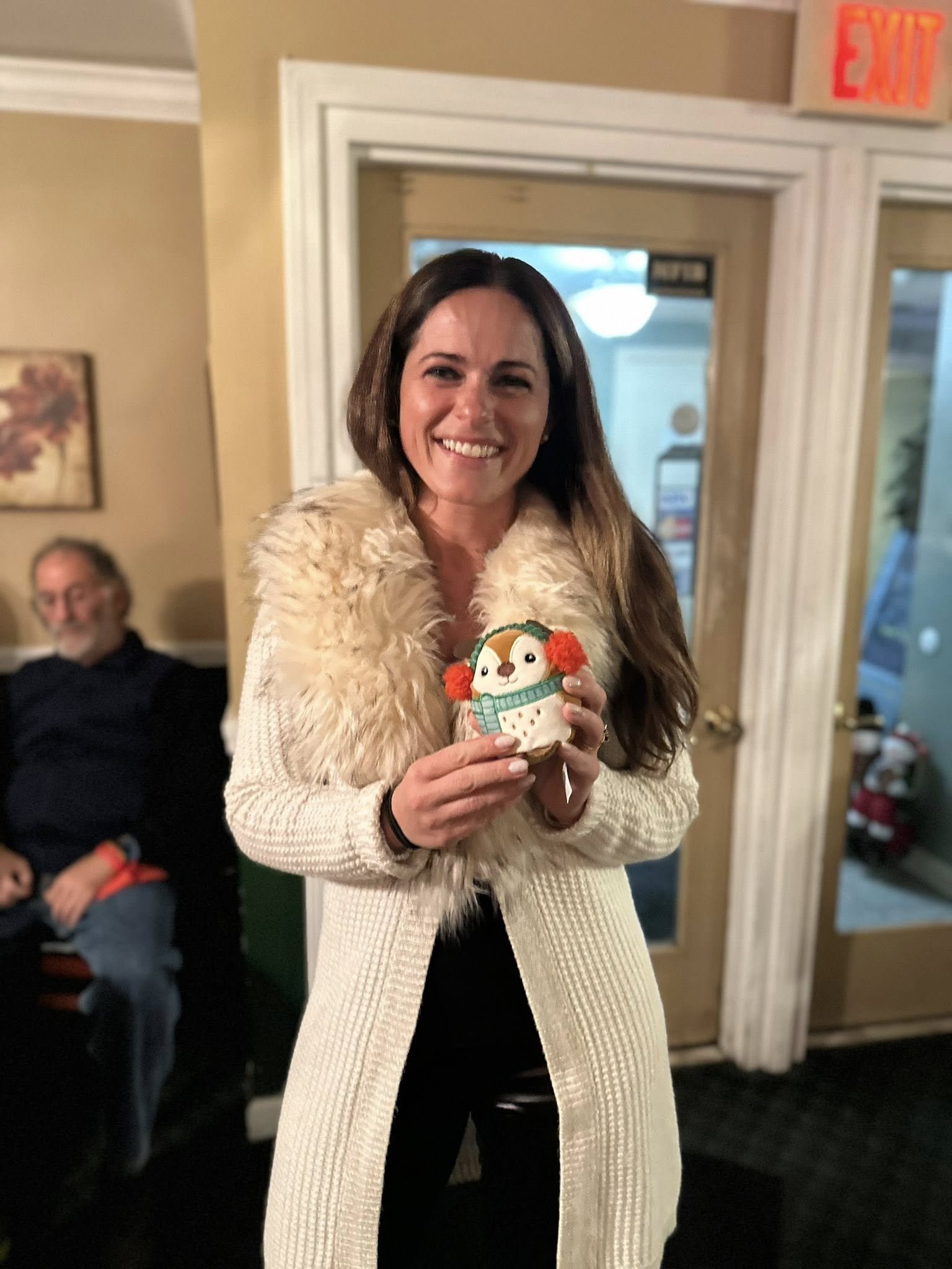 A woman smiling and holding an award in a cozy indoor setting with a man sitting in the background.