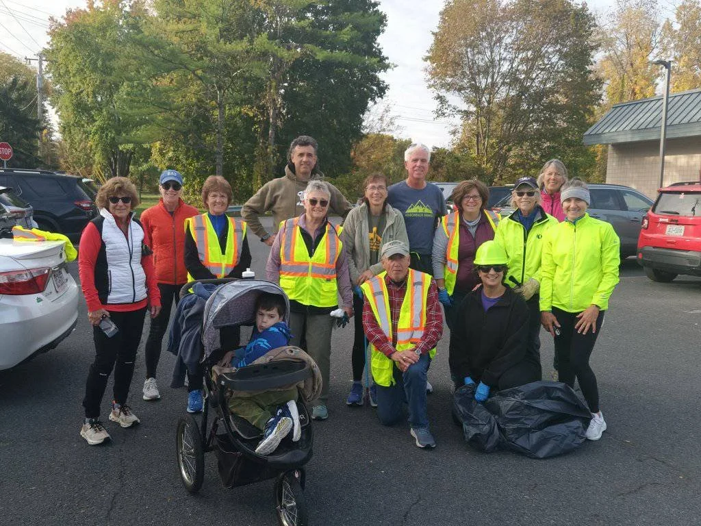 Group of volunteers, mostly women, wearing high-visibility vests and outdoor clothing on a parking lot during daytime, with trees and cars in the background, and a child in a stroller.