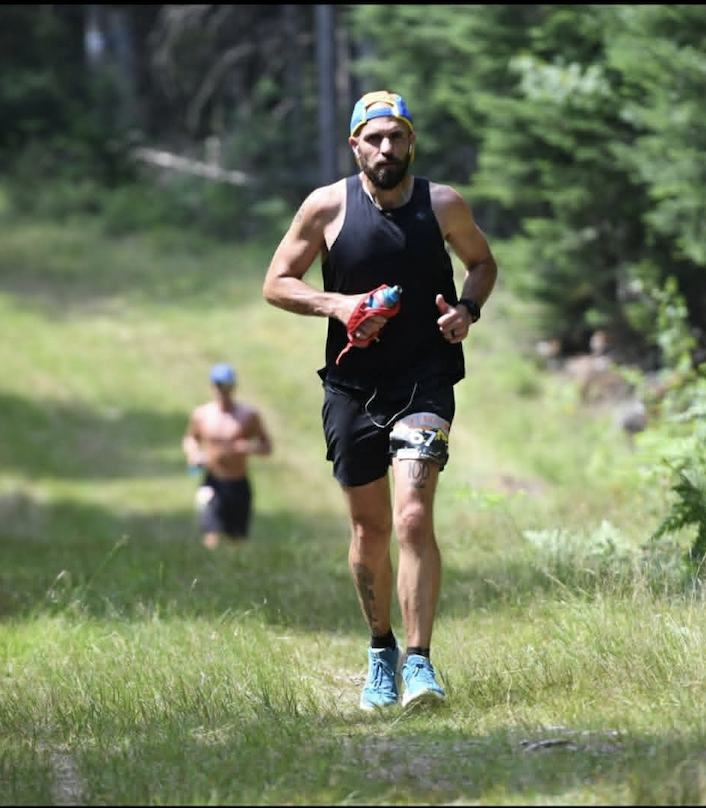 Male runner with a blue cap, sleeveless black shirt, and black shorts running on a grassy trail in a wooded area during a race.