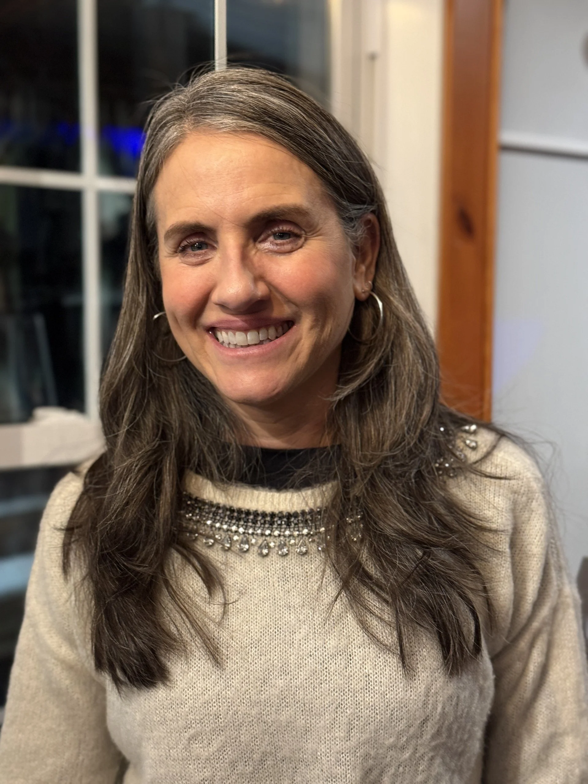 A woman with long brown hair, smiling, wearing a beige sweater with embellishments on the collar, standing indoors near a window.