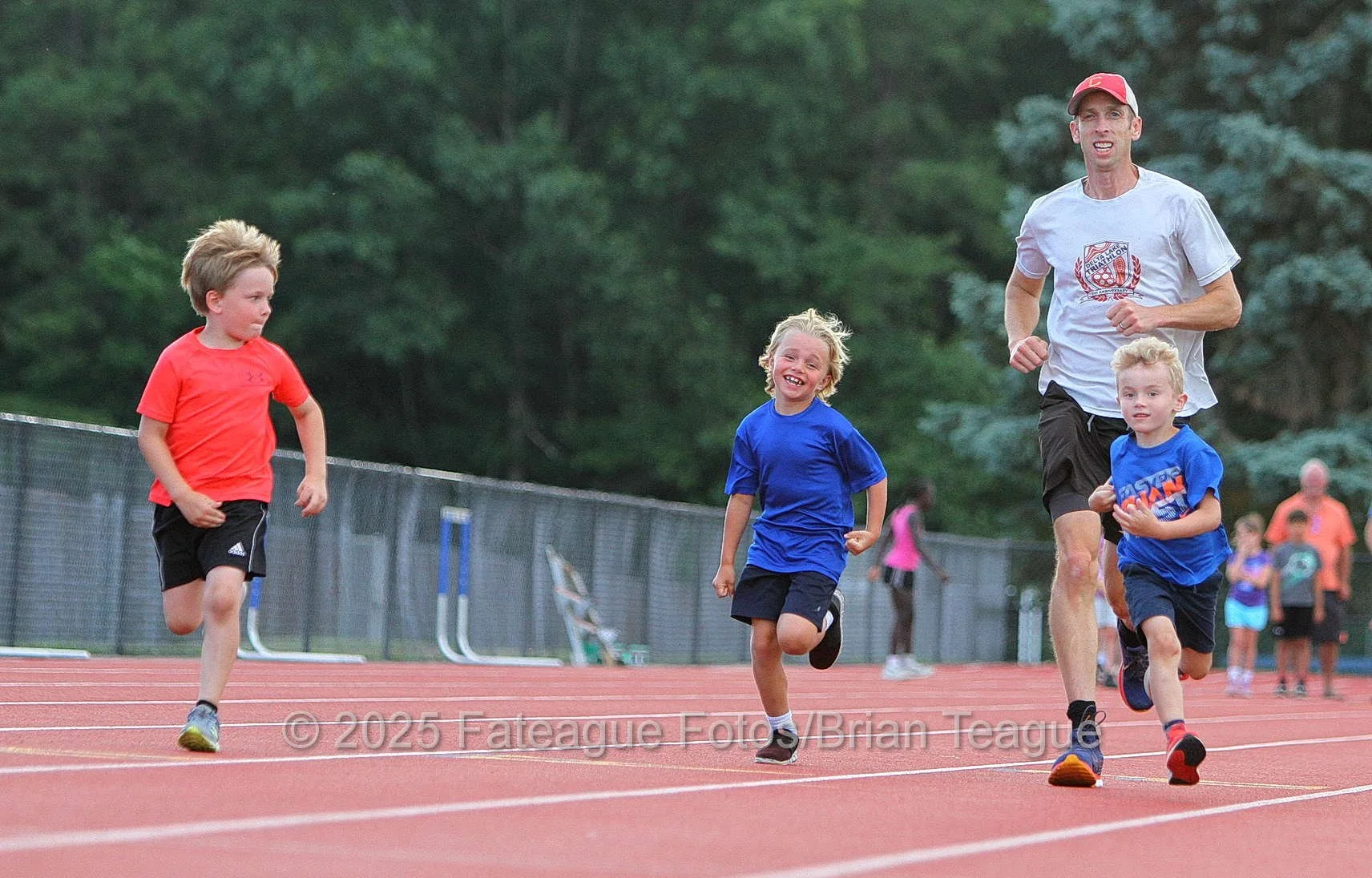 A man and three children running on a track at a sports field, with trees in the background.